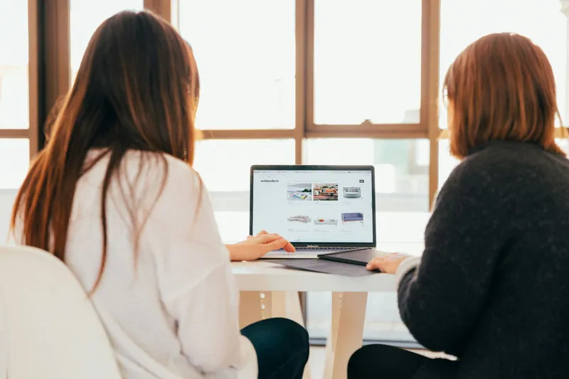 Two colleagues working together at a laptop by a bright window